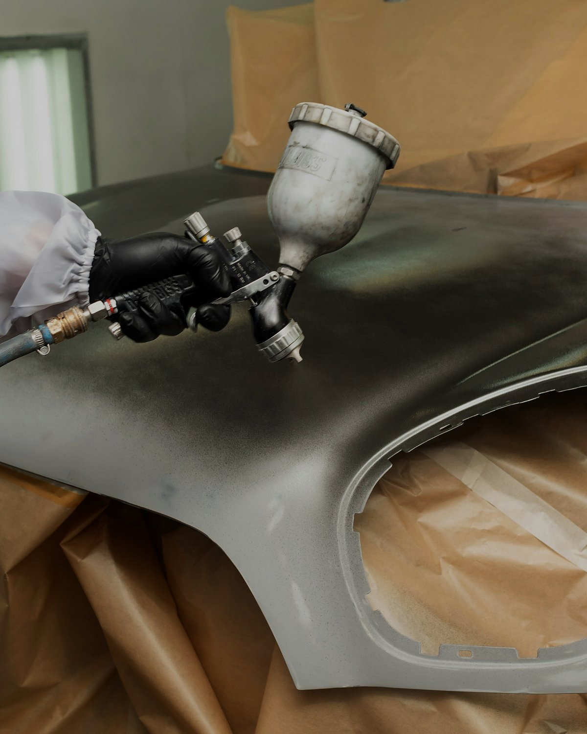 A hand in a black glove and white protective sleeve holding a spray gun over a silver car fender, with brown masking paper covering the surroundings