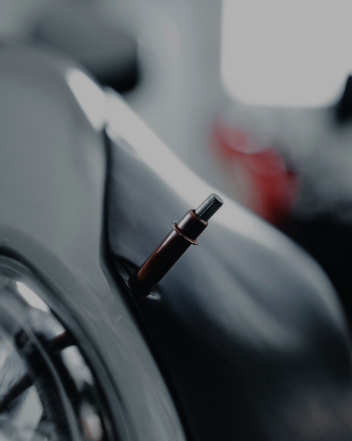 Close-up of a copper-coloured pull stud welded into a dent on a grey car fender next to the headlight, with a blurred workshop in the background