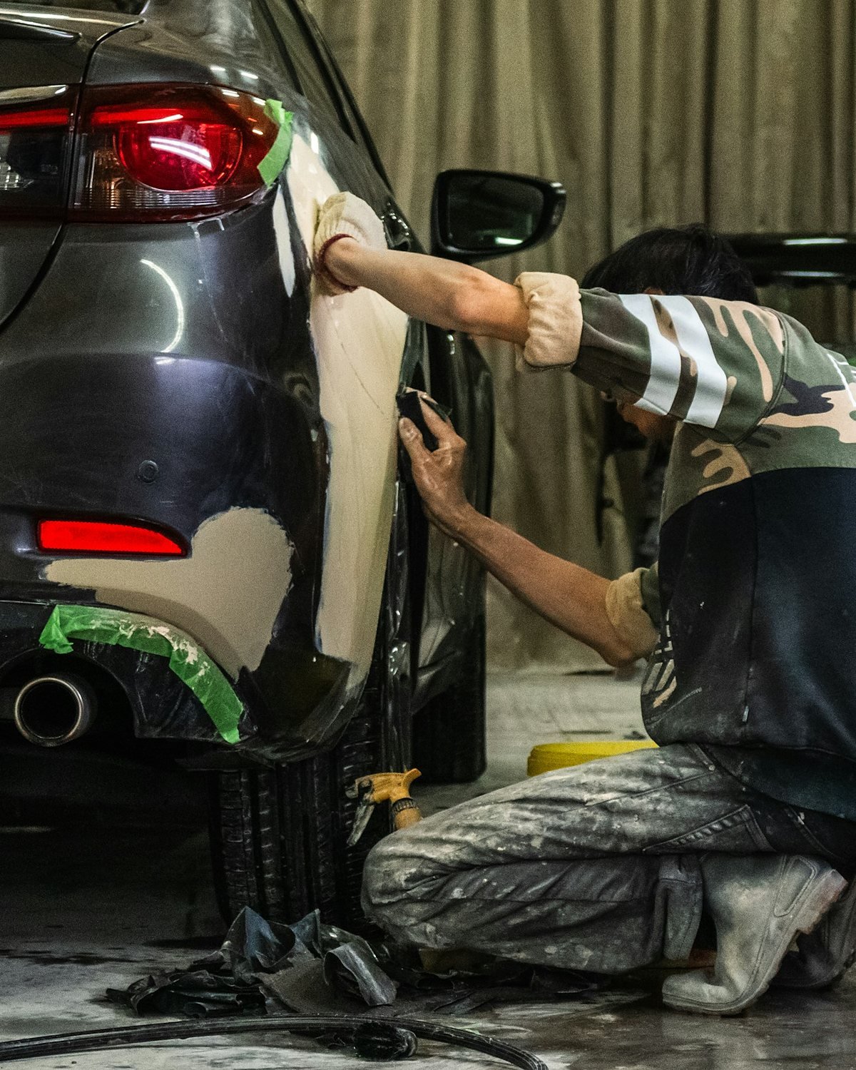A kneeling worker hand sanding patches of pale body filler on the rear quarter panel and bumper of a dark grey car in a workshop
