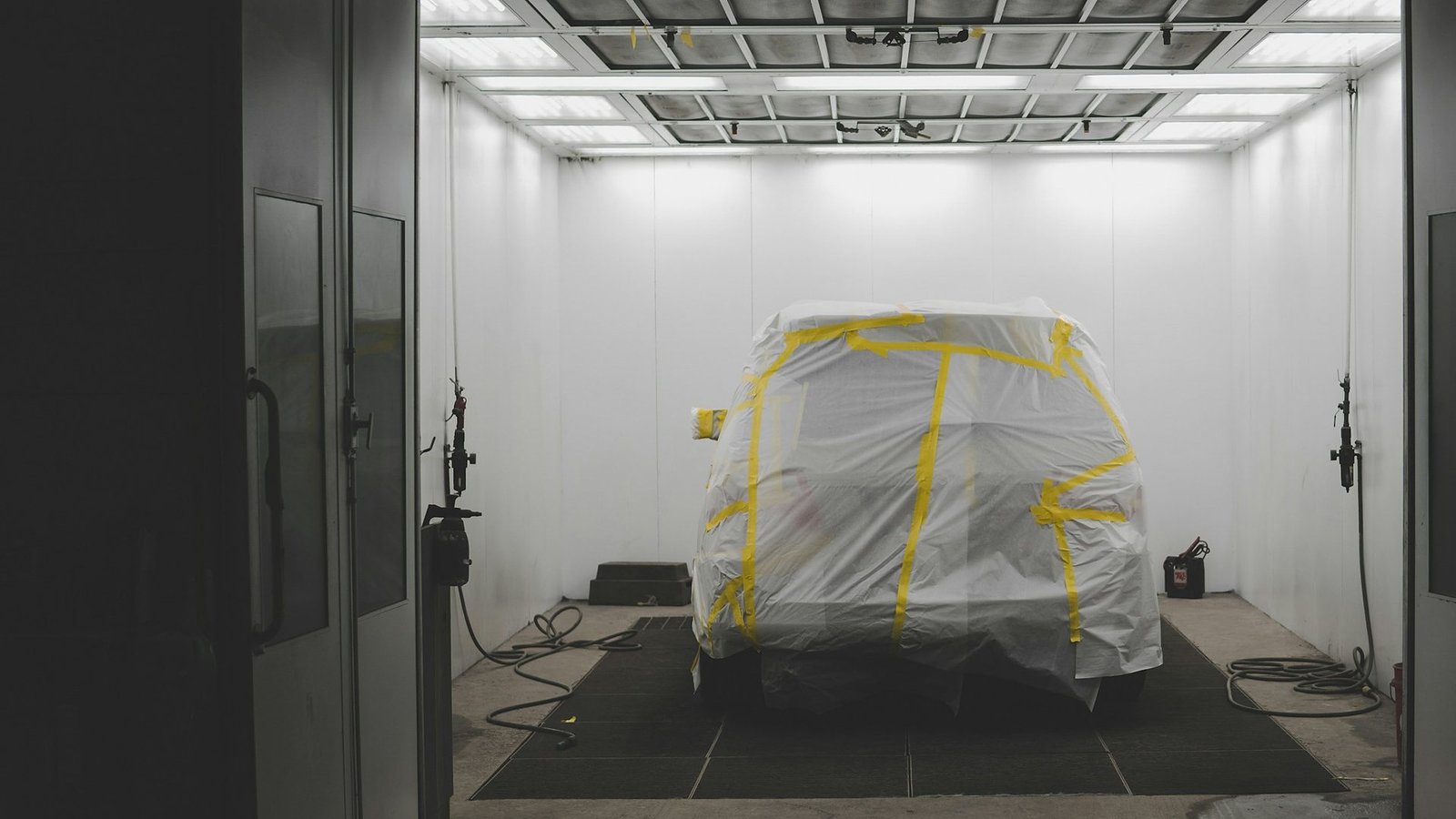 A car covered in plastic sheeting and yellow masking tape inside a white spray booth