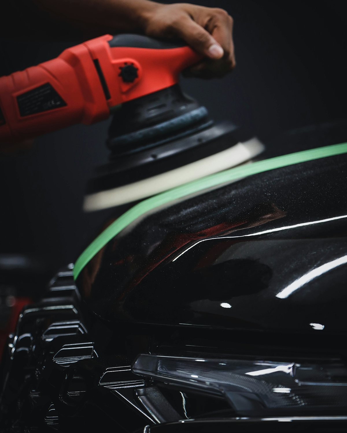 Hand holding a red machine polisher with a foam pad on a black car bonnet edged with green masking tape