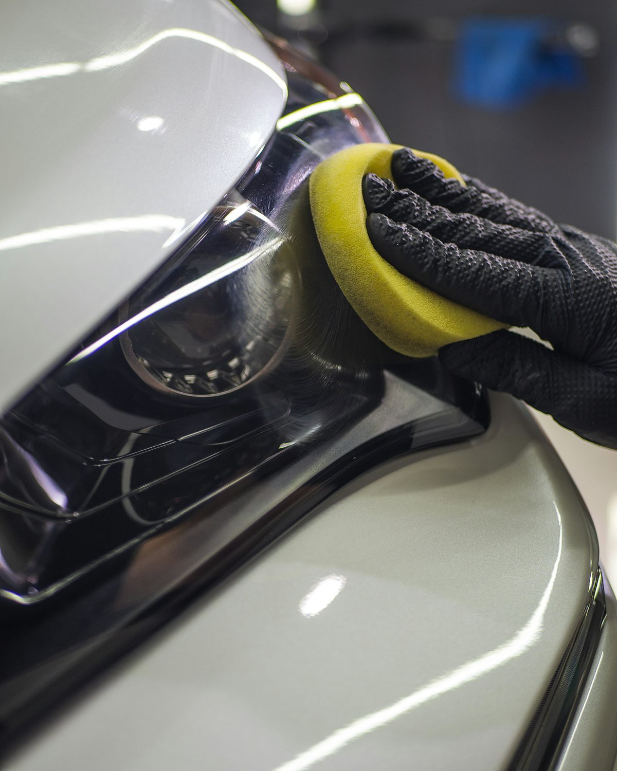 Gloved hand pressing a yellow foam applicator pad onto the headlight lens of a silver car