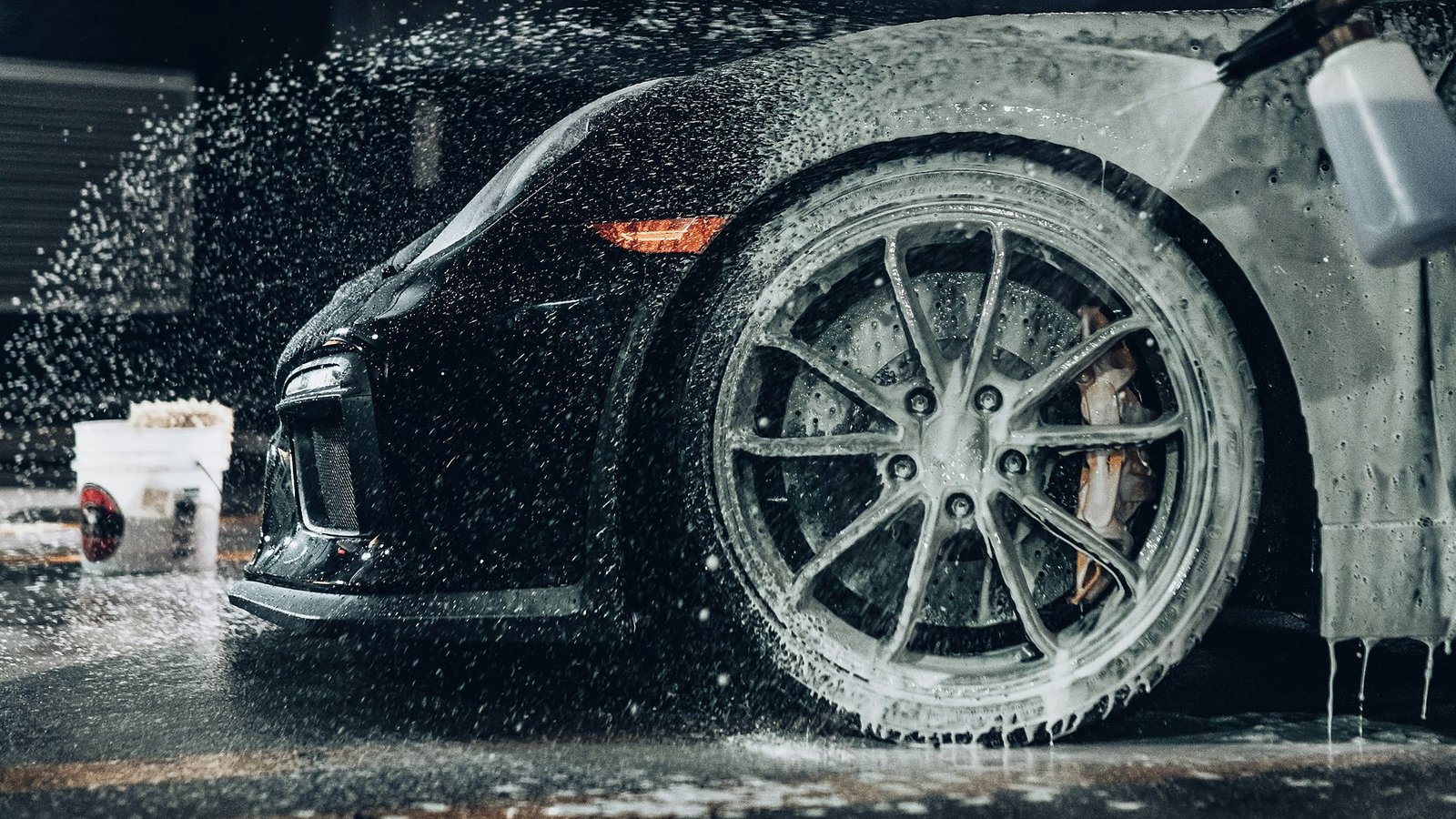 Front wheel of a black sports car under foam and spraying water, with a wash bucket behind it