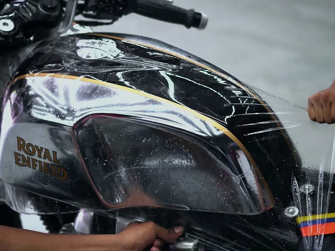 Hands stretching clear film over the chrome tank of a Royal Enfield Continental GT 650.