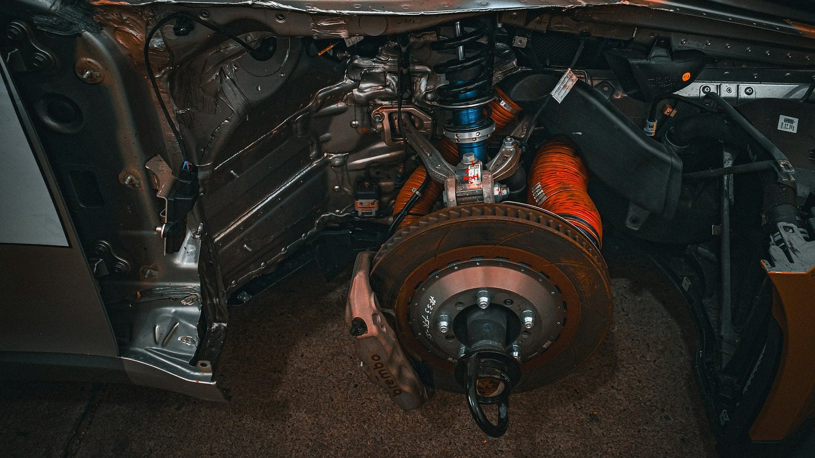 Front corner of a stripped car with the wheel off, showing the brake disc, caliper, a blue coilover and orange ducting
