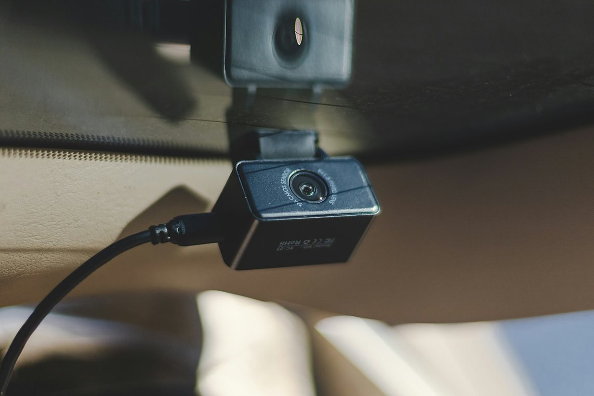 Black dash camera hanging from a mount stuck to car glass, its power cable curving in from the lower left