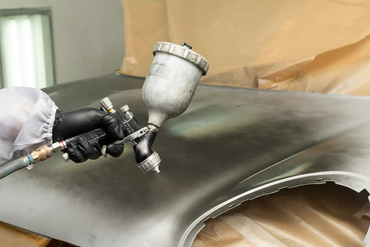 A gloved hand spraying silver paint onto a car fender with a gravity-feed spray gun, brown masking paper behind the panel