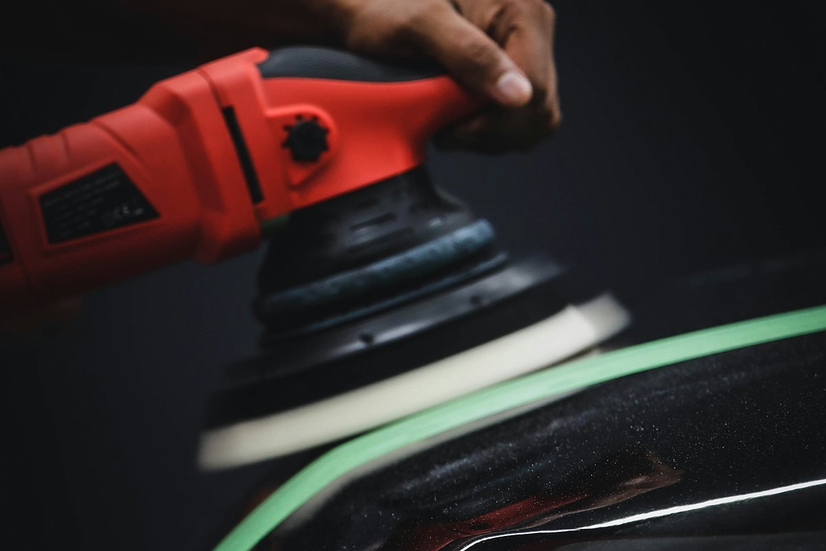 Hand running a red machine polisher with a white foam pad along green masking tape on a black car panel