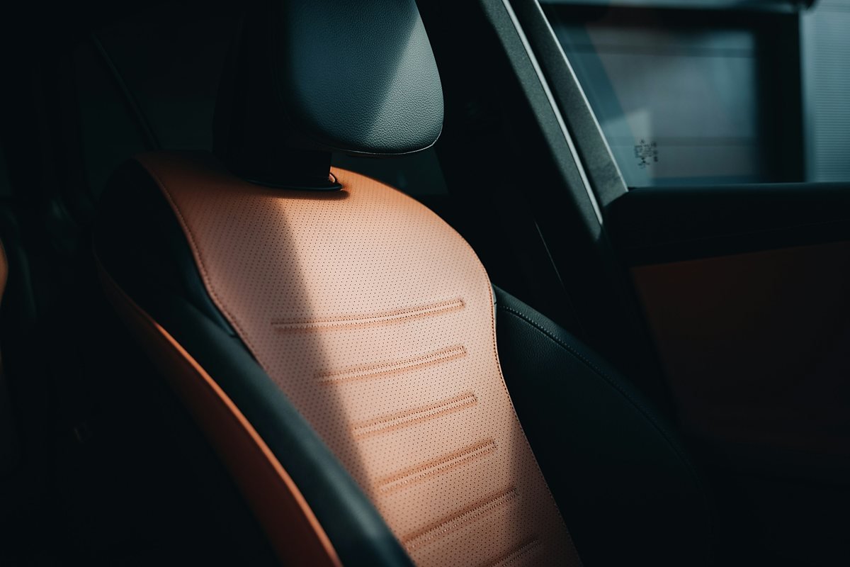 Close-up of a car seat back upholstered in tan perforated leather with stitched horizontal channels, framed by black leather side bolsters and a black headrest, lit by a single shaft of soft light in a dark cabin