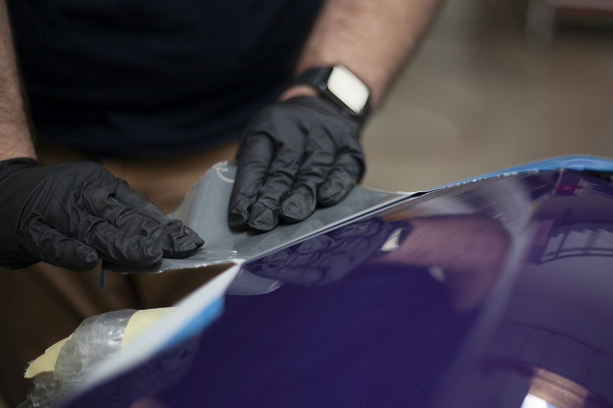 Gloved hands smoothing a sheet of vinyl film onto a glossy dark car panel, with masking tape along the edge